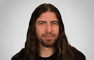 A man with long brown hair and a beard looking forward against a light gray background.