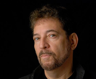 A man with curly brown hair and a salt-and-pepper beard looking toward the camera against a black background.
