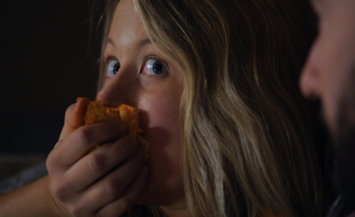 A blonde woman with wide eyes holds a piece of food to her mouth while looking toward a man.