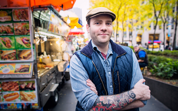 A man with tattoos on his arm stands with crossed arms in front of a street food truck.