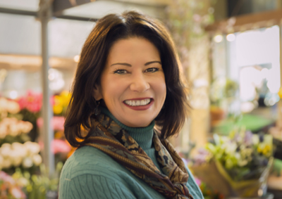 Smiling woman with dark hair wearing a teal turtleneck and patterned scarf in a flower shop.
