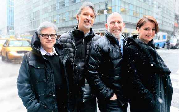 Four smiling people in dark winter coats standing on a snowy city street with a yellow taxi in the background.