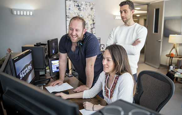 A man and woman look at a computer monitor displaying the word 'Scraps' while a man stands behind them.