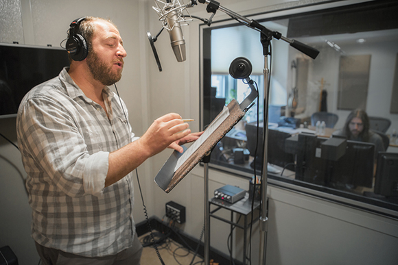 A bearded man wearing headphones sings into a microphone while reading from a script in a recording studio.