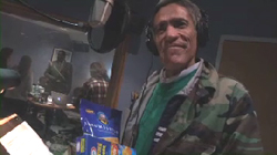 A man in a camouflage jacket and headphones smiles in a dimly lit room with snacks in the foreground.