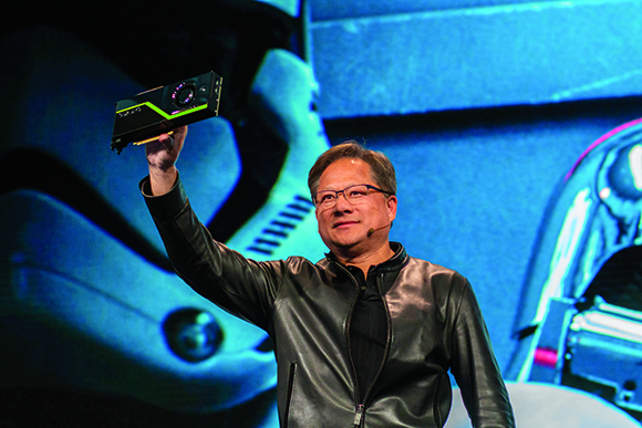 A man in a black leather jacket holds a small black rectangular device aloft during a presentation.