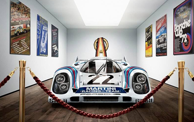 A white Porsche race car with blue and red stripes and the number 22 is displayed in a gallery with racing posters on the wal