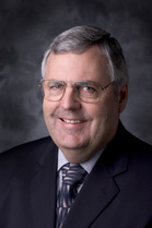 A man with graying hair and glasses wearing a dark suit, light shirt, and patterned tie.