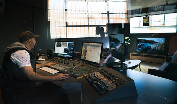 A man sits at a desk with multiple monitors and a large mixing console in a dark studio.