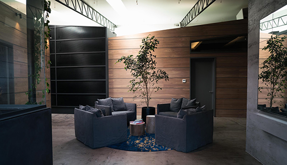 A lounge with dark grey sofas, a small tree in a pot, and wood-paneled walls.