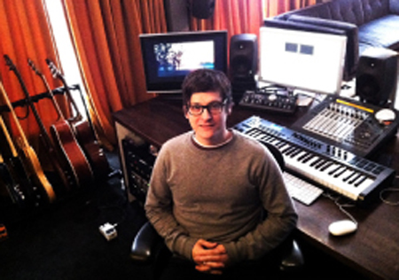 A man with glasses sits at a desk in a music studio filled with monitors, keyboards, and guitars.