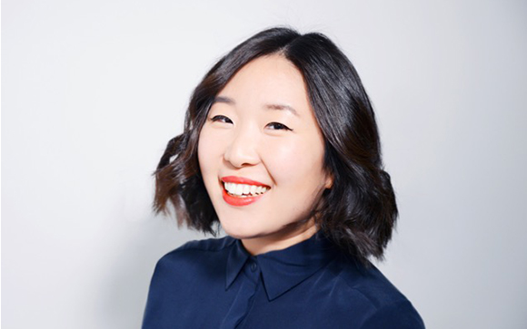 A smiling woman with wavy dark hair and red lipstick wearing a dark blue collared shirt.