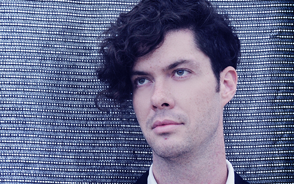A man with dark curly hair looks off to the side against a blue and white textured backdrop.