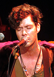 A man with curly hair singing into a microphone with his eyes closed.