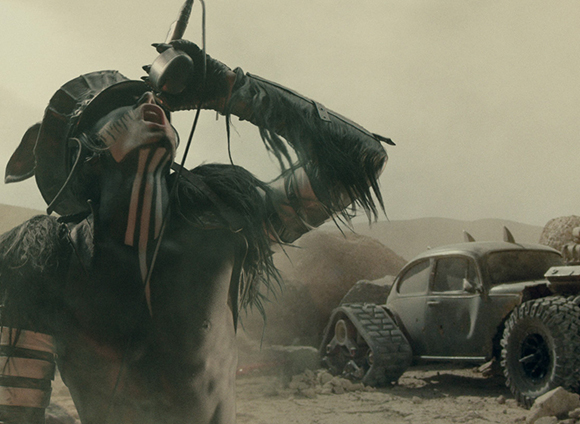 A person in dark, fringed armor tilts their head back to drink from a small cup in a desert landscape.