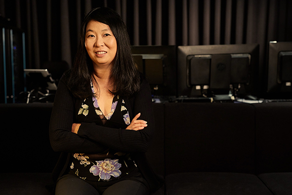 A woman with long dark hair and a floral top sits with her arms crossed in a dark studio.