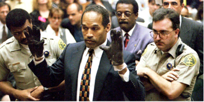 A man in a dark suit and patterned tie raises his hands while surrounded by people and security officers.