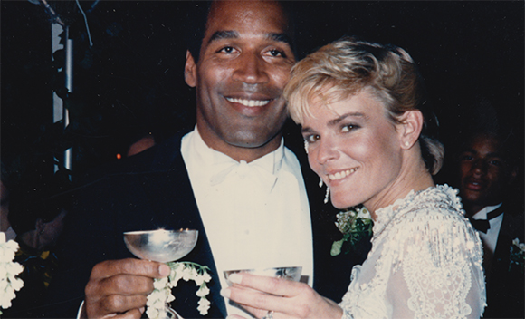 A man in a tuxedo and a woman in a white lace dress smile while holding glasses.