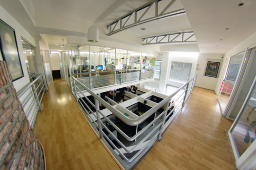 High-angle view of a two-story modern office with light wood floors, glass walls, and metal railings.