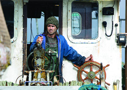 A man in a blue jacket and knit cap stands on a boat near a large wooden steering wheel.
