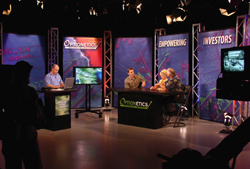 People sit at desks in a television studio with large screens and colorful backdrops.