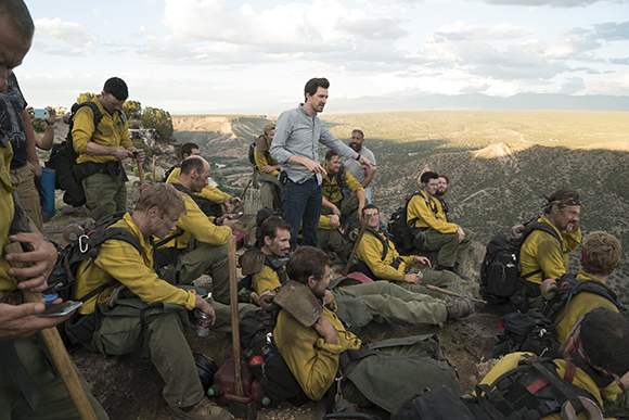 A group of people in yellow uniforms sit and stand on a rocky ledge overlooking a wide valley under a cloudy sky.