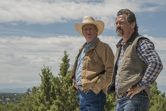 Two men wearing cowboy hats and vests stand outdoors against a blue sky with clouds.