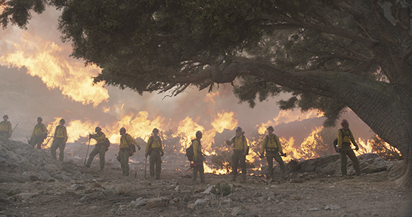 Wildfire firefighters in protective gear stand on a rocky slope as flames burn in the background.