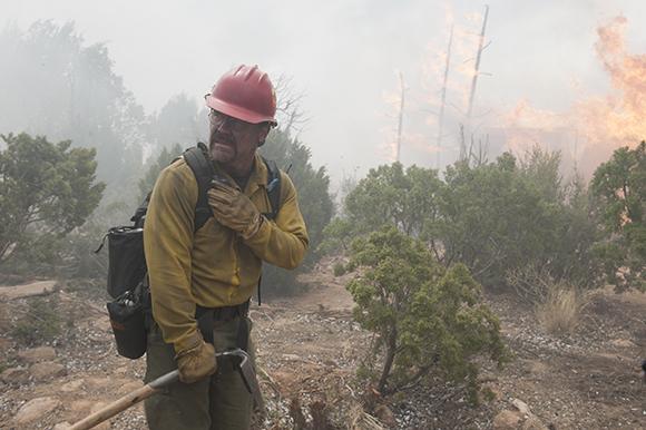 A firefighter in a red helmet and yellow shirt stands in a smoky landscape with flames in the background.