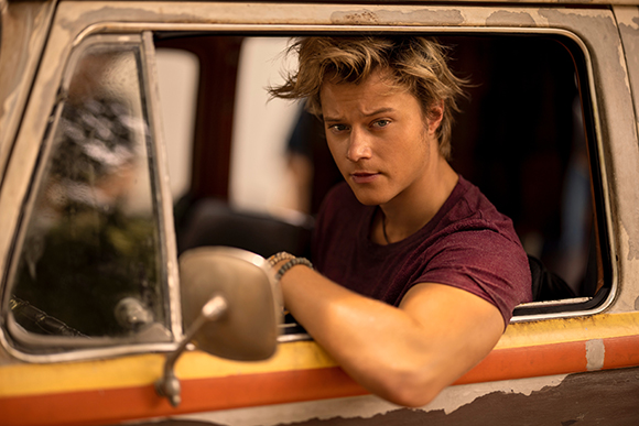 A young man with blond hair leans his arm out of a vintage truck window and looks forward.