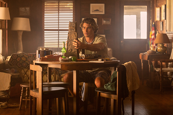 A man in a light t-shirt sits at a round wooden table in a dimly lit room, holding a tall, dark textured object.
