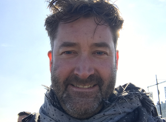 Close-up selfie of a smiling man with brown hair and a beard wearing a grey patterned scarf.