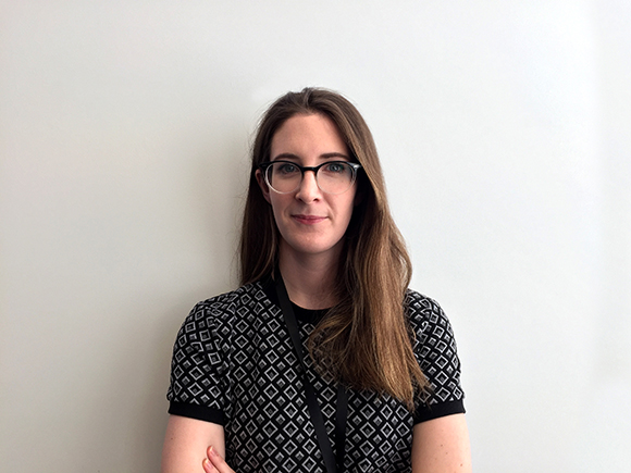 A woman with long brown hair and black-rimmed glasses wearing a black and white patterned shirt against a white wall.
