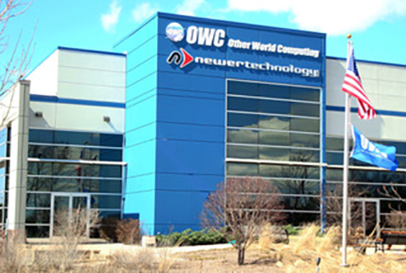 A blue and silver commercial building with OWC and Newer Technology logos and two flags flying.