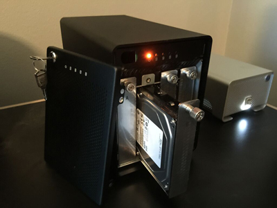 A black NAS device with its front panel removed, revealing an internal hard drive and a glowing orange status light.