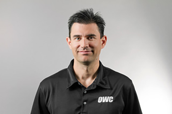A man with short dark hair wearing a black polo shirt with an OWC logo, smiling against a gray background.