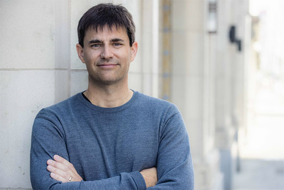 A man with dark hair and a blue sweater stands with arms crossed against a light-colored stone wall.