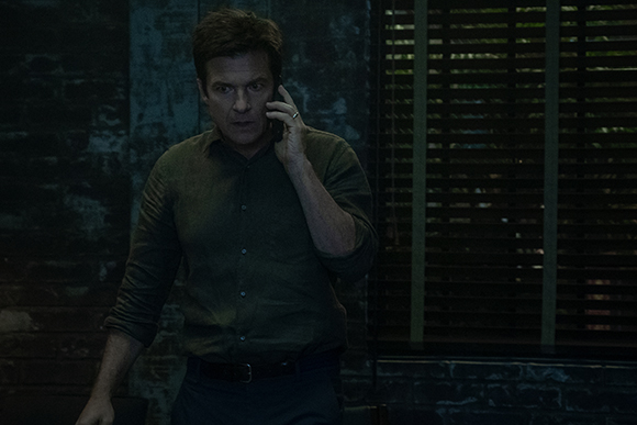 A man in a dark green button-down shirt talks on a mobile phone in a dimly lit room.