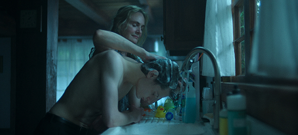 A woman stands behind a shirtless boy, washing his hair at a bathroom sink in dim lighting.