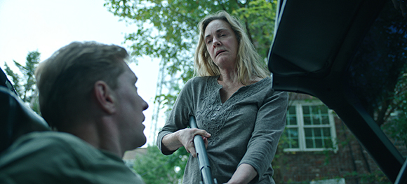 A woman in a grey shirt looks down at a man sitting in a car outdoors.