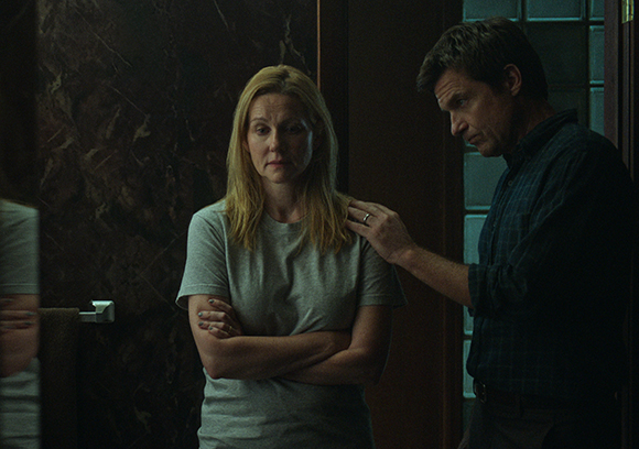 A man places his hand on the shoulder of a blonde woman standing with her arms crossed in a dimly lit room.