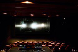 A dark movie theater showing a bright, glowing scene on a large screen above rows of red seats.