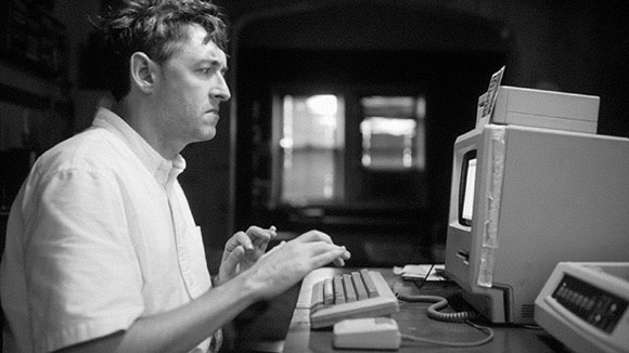 Black and white photo of a man typing on a vintage computer in a dimly lit room.