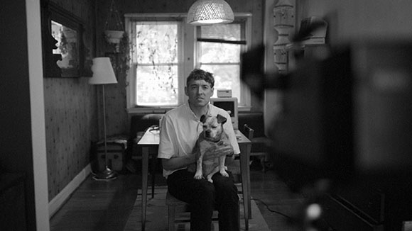 Black and white photo of a man sitting at a table holding a small dog in a room with a window.