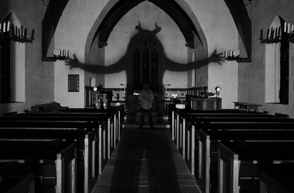 Black and white photo of a large, dark shadow with outstretched arms cast on a church wall above a person.