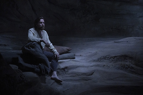 A man with a beard sits on the ground in a dark, rocky cave, leaning against a makeshift bed.