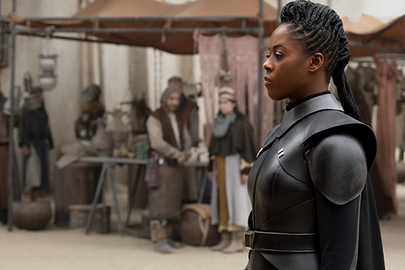 A woman with braided hair wearing black leather armor stands in a blurred outdoor market.