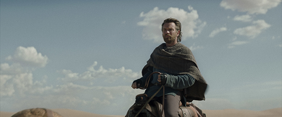 A bearded man in a dark cloak riding a horse through a vast desert under a cloudy sky.