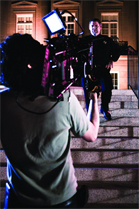 A person holds a professional video camera, filming a man walking down stone stairs at night.
