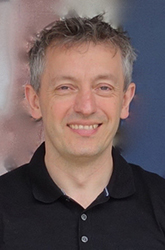 A smiling man with short, wavy grey hair wearing a black polo shirt.